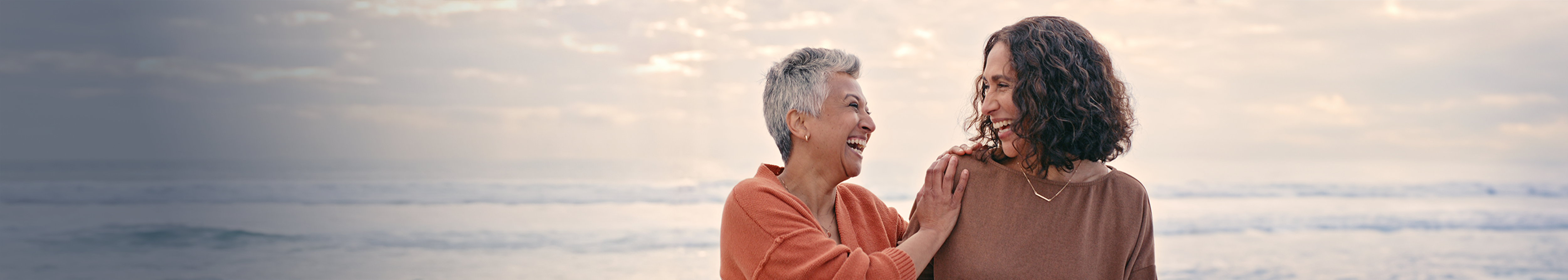 Two diverse women laughing at the beach, possibly discussing a breast screen appointment.